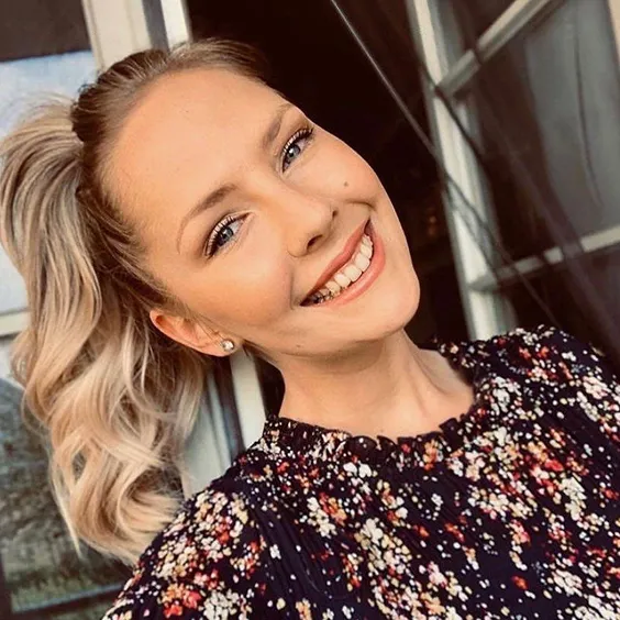 Smiling young woman with blonde hair in a ponytail, wearing a black floral top.