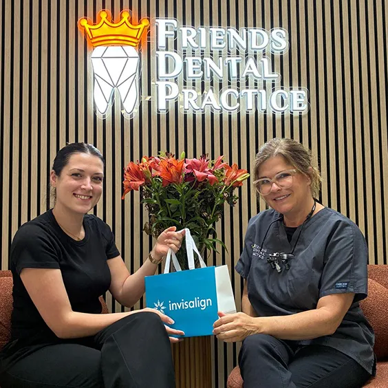 Two women smiling inside Friends Dental Practice, one holding an Invisalign bag, with a flower arrangement and a wooden slat wall in the background.