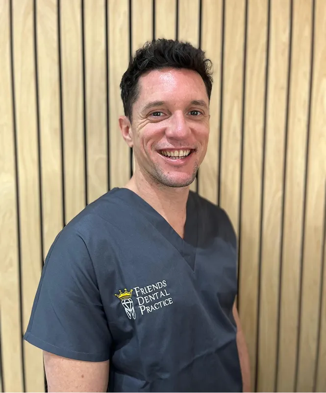 Smiling man wearing a gray scrub top with Friends Dental Practice logo, standing against a wood-paneled wall.