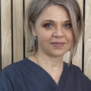 Portrait of a woman with short blonde hair wearing a dark blue scrub top and earrings, standing in front of a wooden panel background.