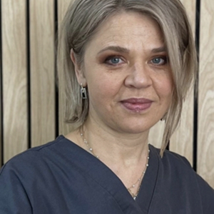 Portrait of a woman with short blonde hair wearing a dark blue scrub top and earrings, standing in front of a wooden panel background.