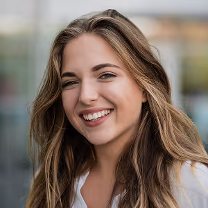 Young woman with long light brown hair smiling outdoors with a blurred background.