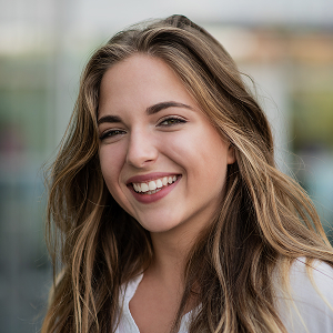 Young woman with long light brown hair smiling outdoors with a blurred background.