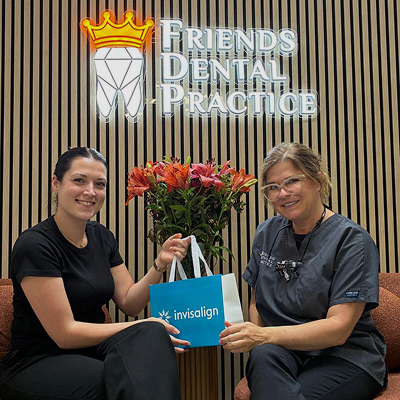 Two women sitting and smiling in front of a Friends Dental Practice sign, holding a blue Invisalign bag.