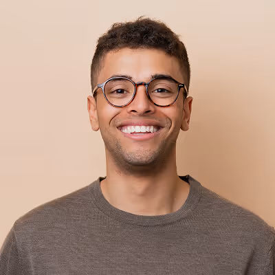 Smiling young man with short curly hair and round glasses wearing a brown sweater against a beige background.