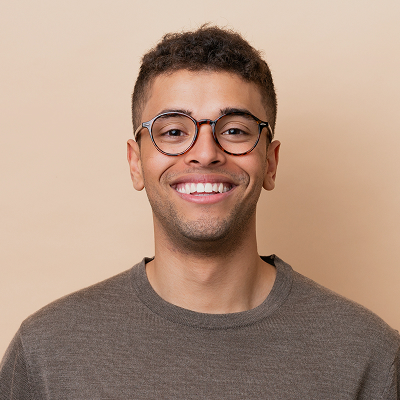 Smiling young man with short curly hair and round glasses wearing a brown sweater against a beige background.