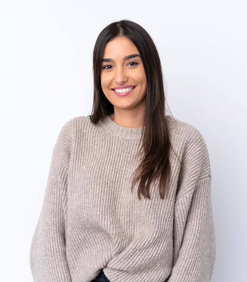 Smiling young woman with long dark hair wearing a beige knit sweater against a white background.