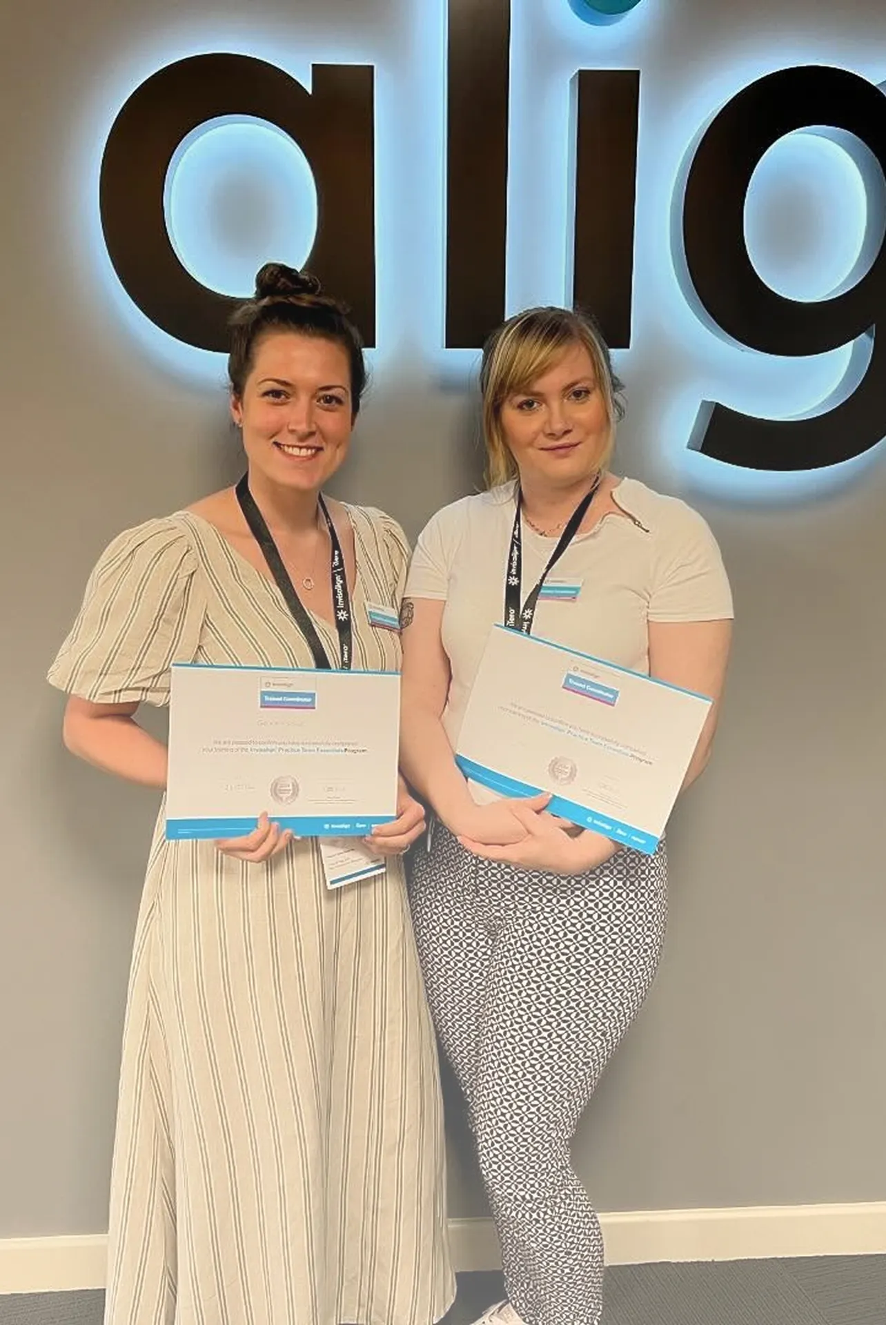 Two women standing in front of a wall with large letters, each holding a certificate and wearing work badges.