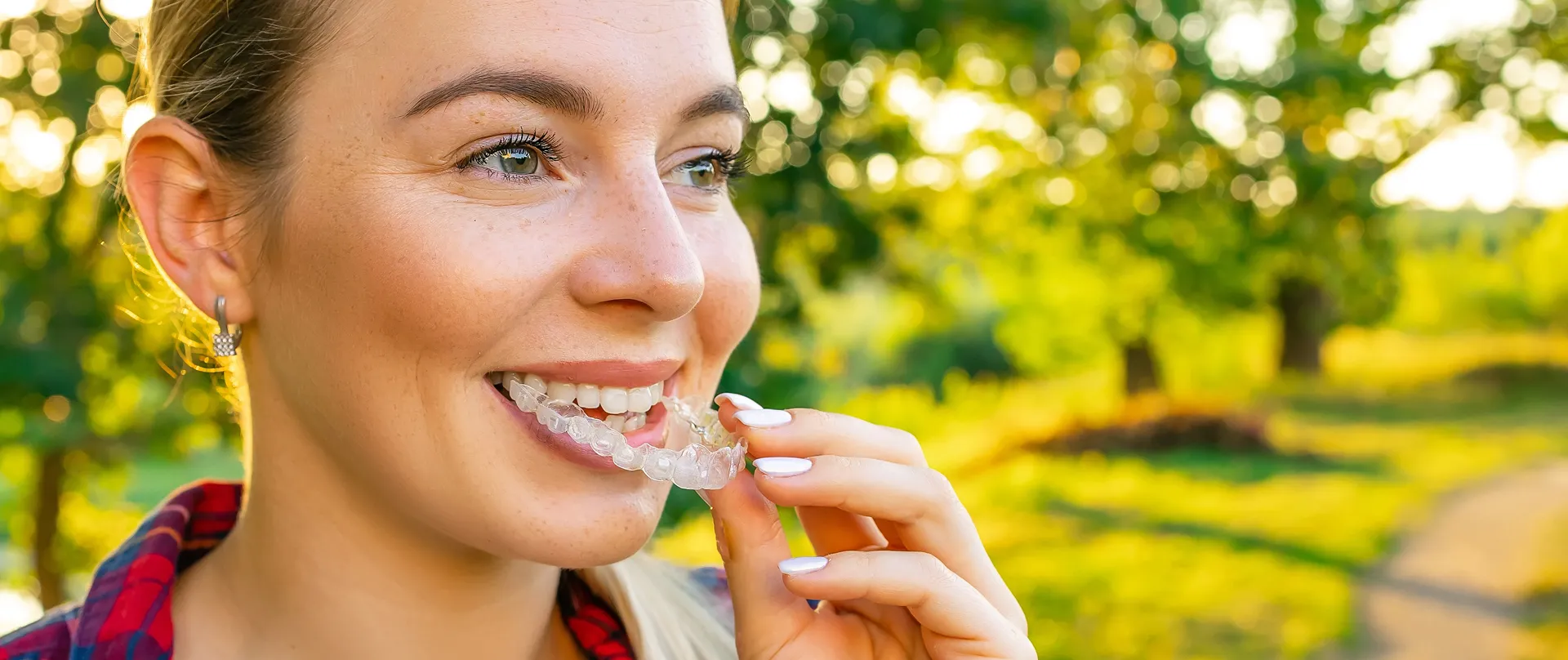 Smiling woman outdoors placing a clear Invisalign aligner on her teeth with a blurred green background.