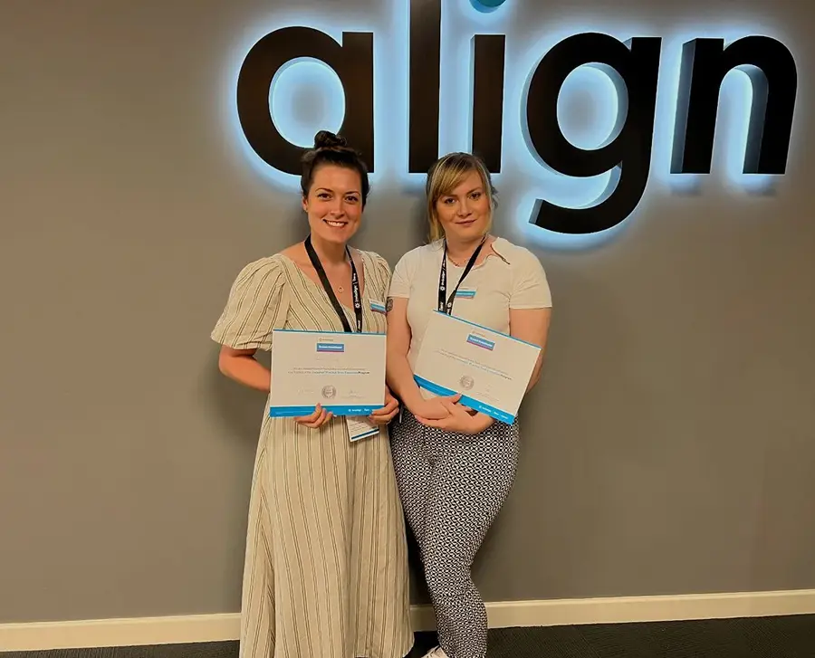 Two women standing and smiling while holding certificates in front of a wall with the illuminated word 'align'.