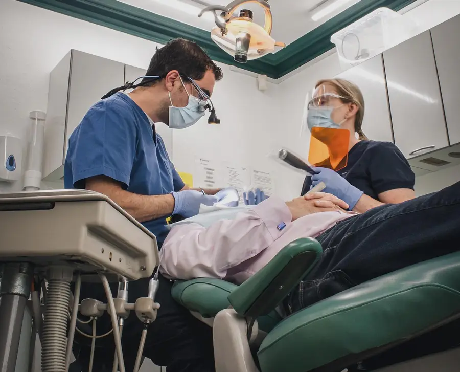 Dentist and assistant wearing masks and gloves performing dental procedure on patient reclining in dental chair.