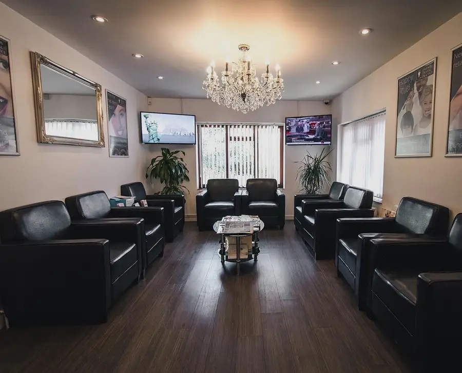 Waiting room with black leather chairs arranged on both sides, a small center table with magazines, two plants, two wall-mounted TVs, and a chandelier ceiling light.