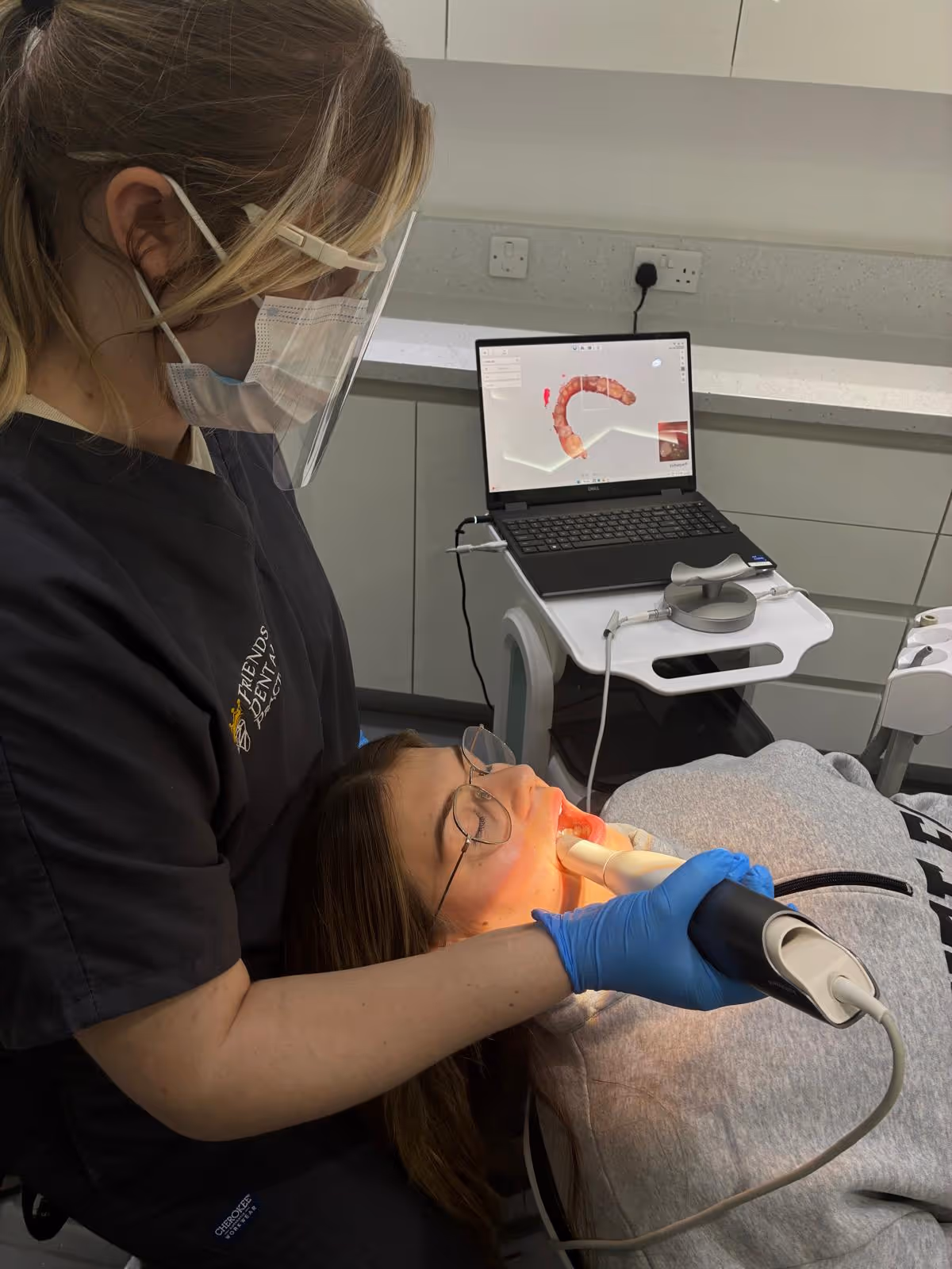Dental professional wearing a face shield and mask using a dental scanner inside a patient's mouth, with a 3D dental image displayed on a laptop.