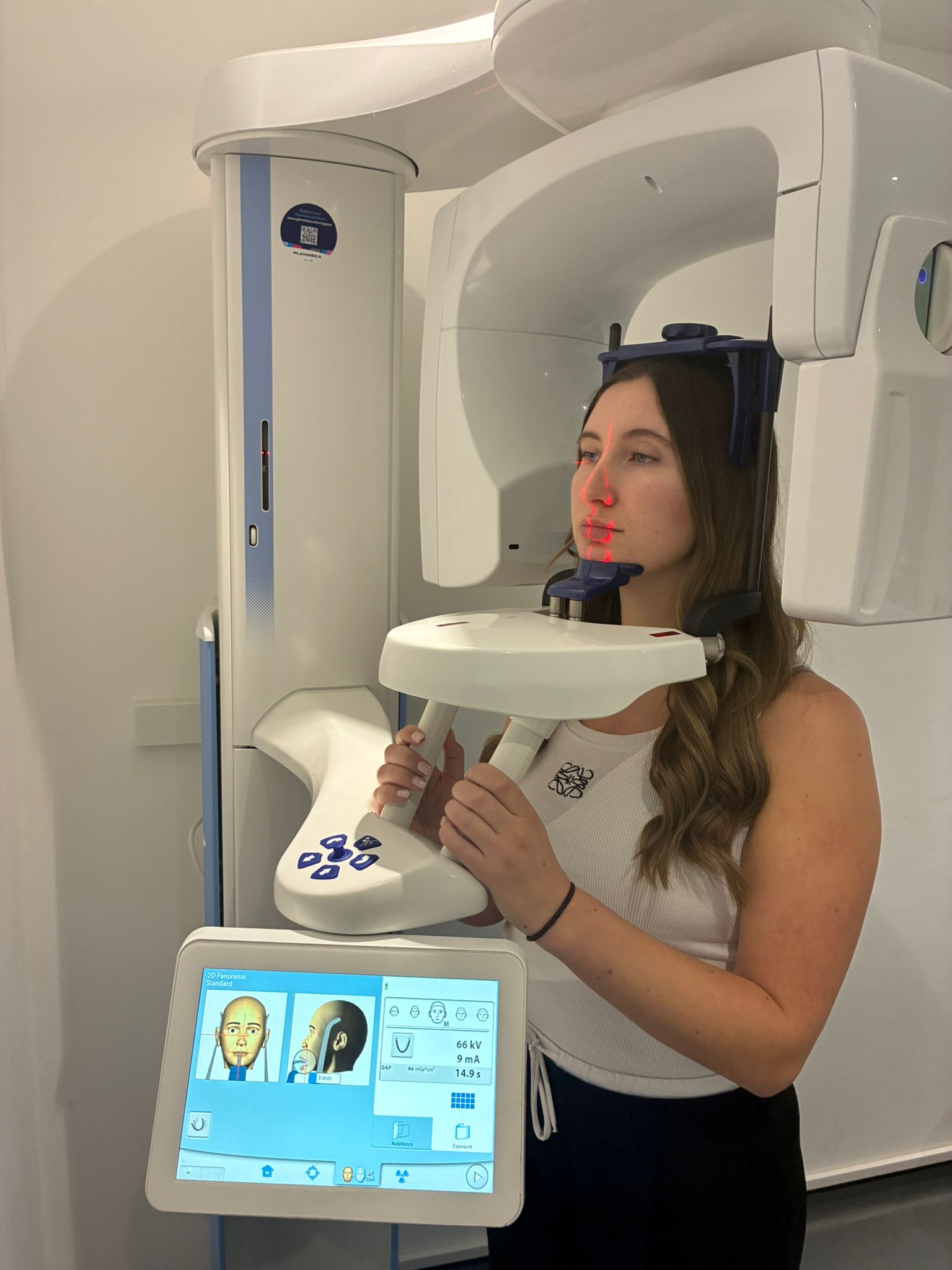 Woman standing in a dental panoramic X-ray machine with red laser guide lines on her face, holding handles, and a control screen displaying head positioning and scan settings.