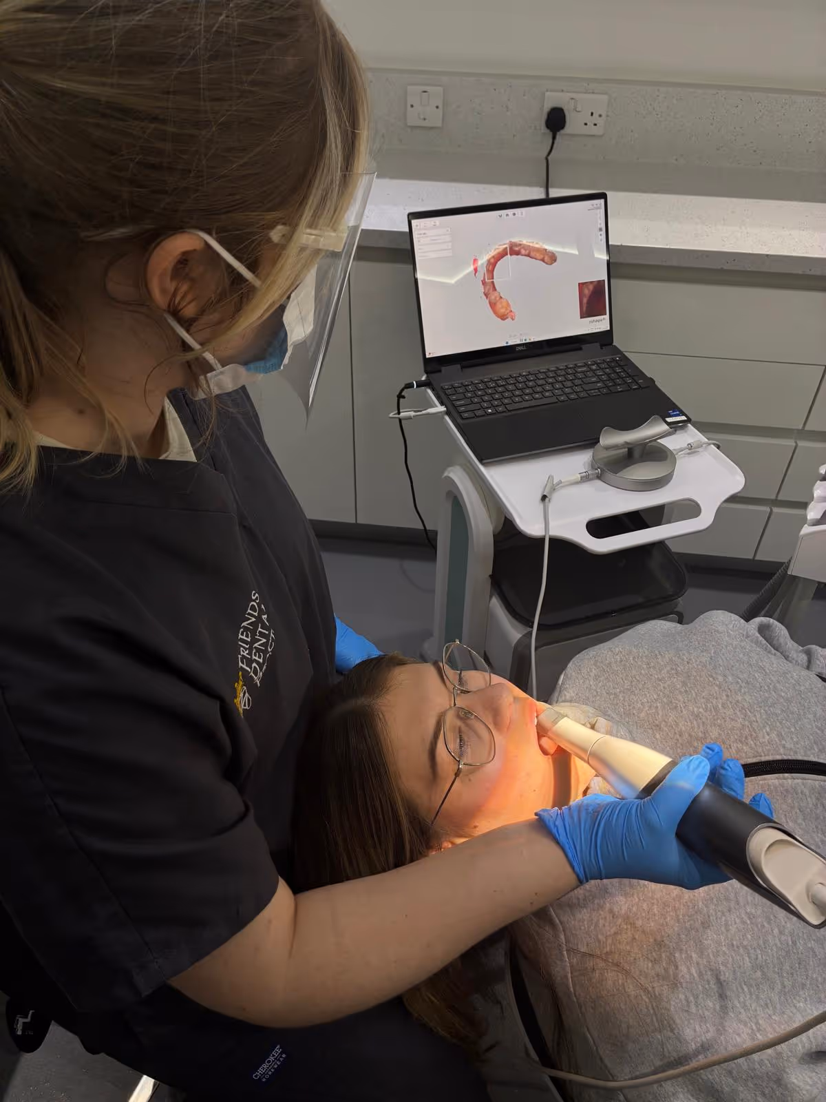 Dental professional wearing protective gear uses an intraoral scanner on a patient lying back with a dental 3D model displayed on a laptop screen nearby.