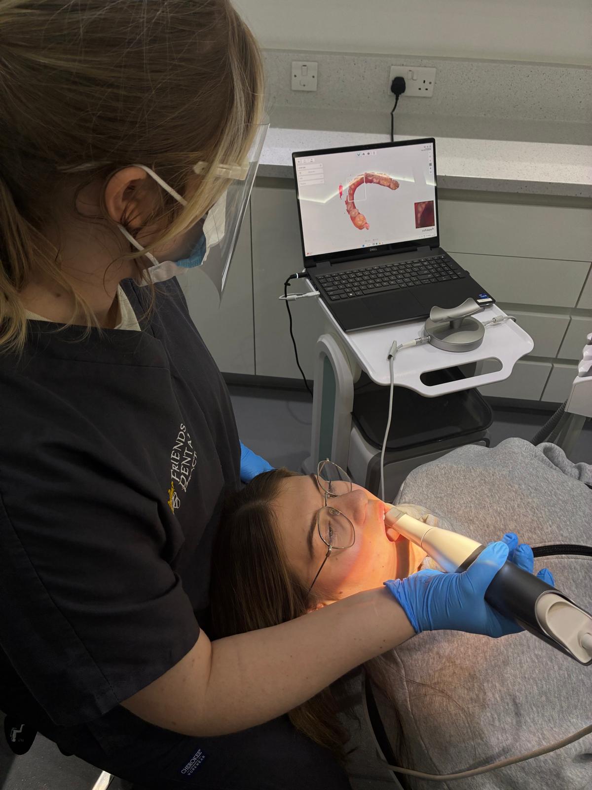 Dental professional wearing protective gear uses an intraoral scanner on a patient lying back with a dental 3D model displayed on a laptop screen nearby.