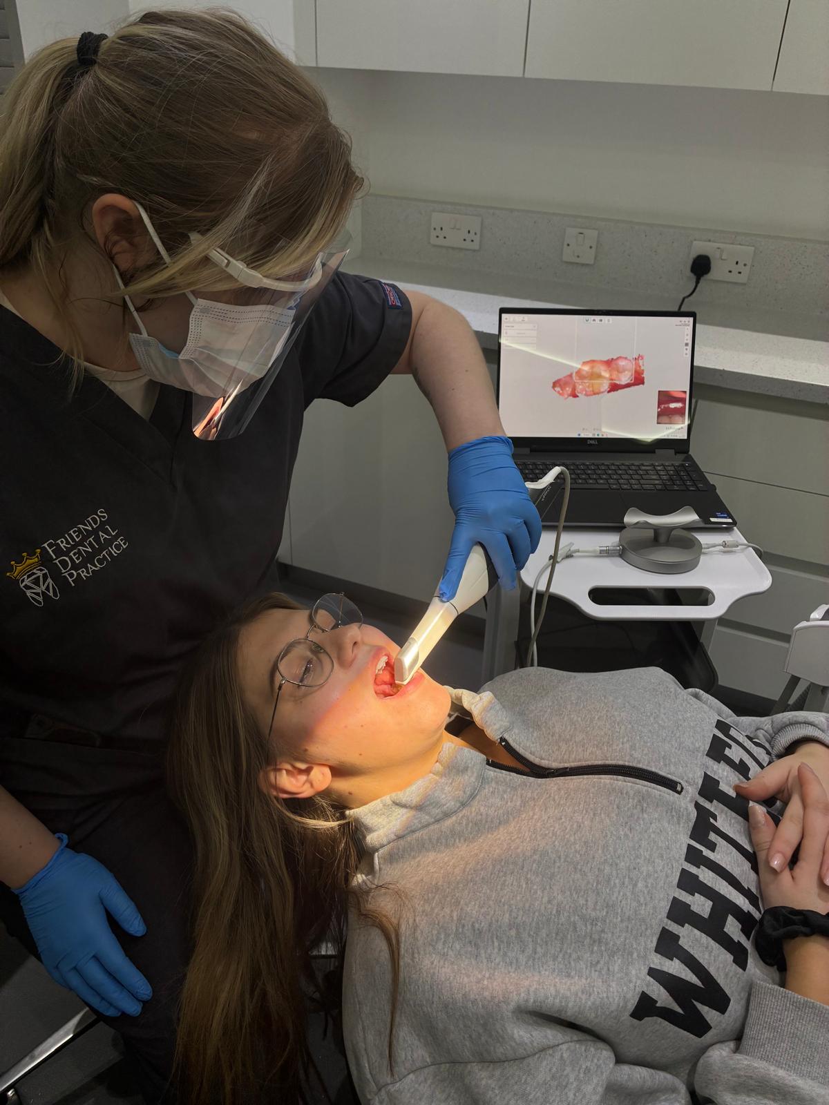 Dentist wearing protective mask, face shield, and gloves using an intraoral scanner inside a patient's mouth with a 3D dental image displayed on a laptop screen.