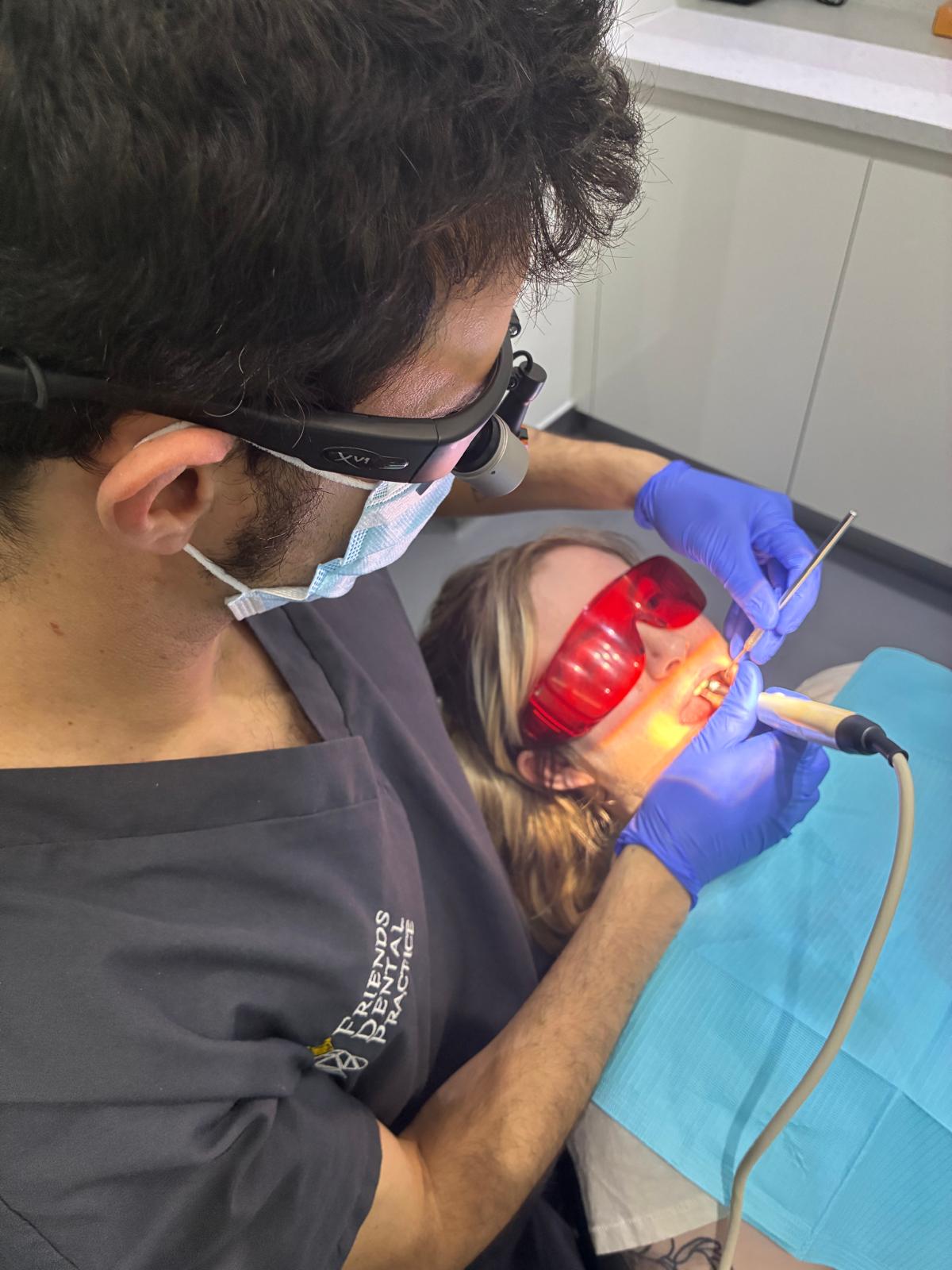 Dentist wearing magnifying glasses and mask performing a dental procedure on a patient wearing red protective glasses.