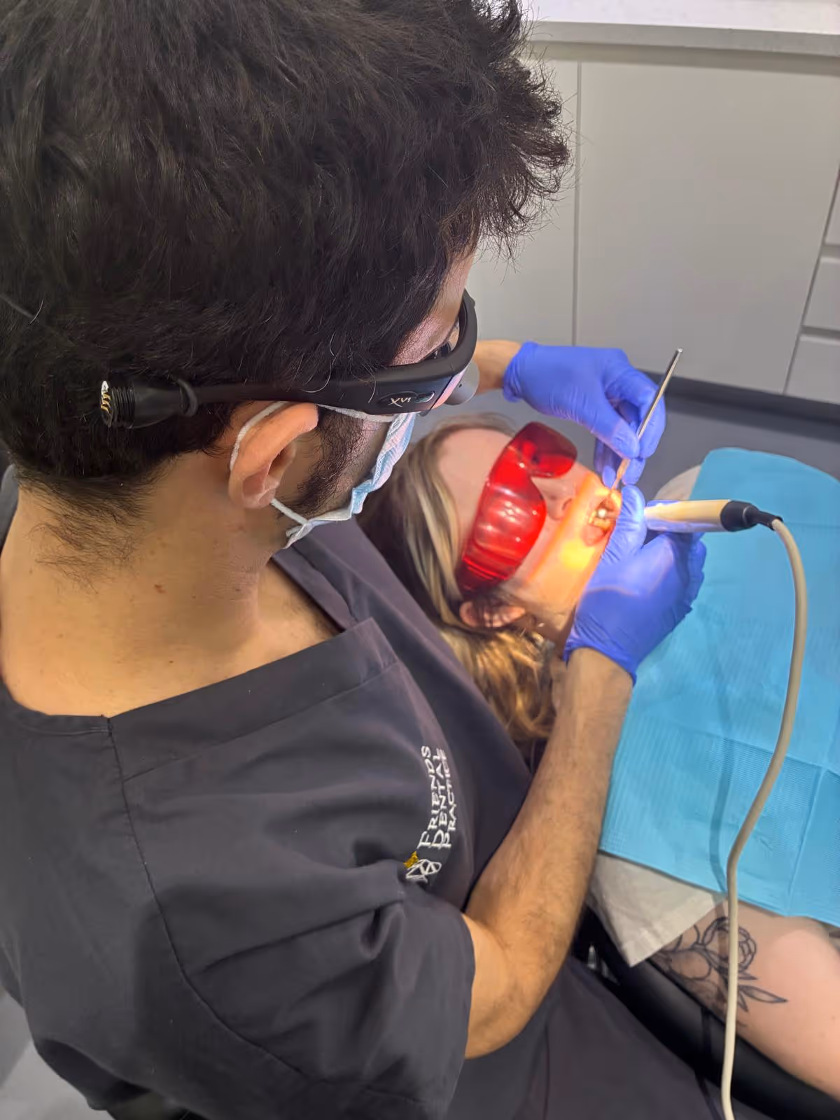 Dentist wearing glasses and mask performing dental treatment on patient lying down with red protective glasses.