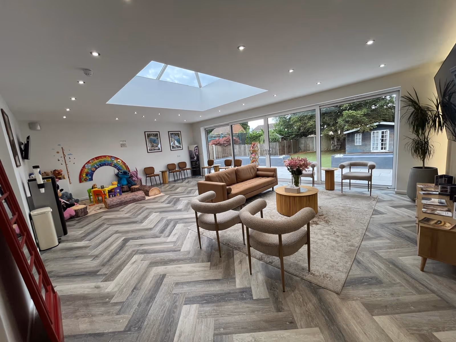Modern waiting room with a brown sofa, beige chairs around a round wooden table, a large skylight, children's play area with toys, and glass doors overlooking a garden with a blue shed.