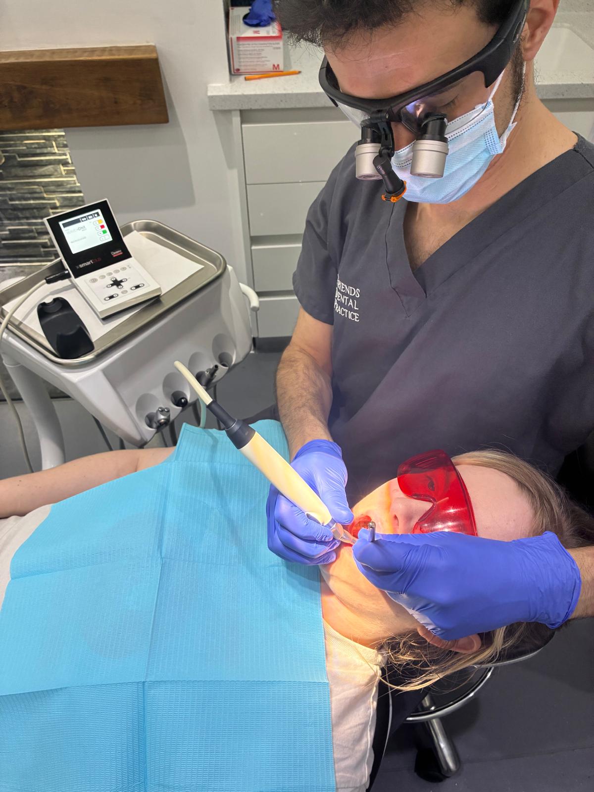 Dentist wearing magnifying glasses and mask performing a dental procedure on a patient with red protective glasses and blue dental bib.