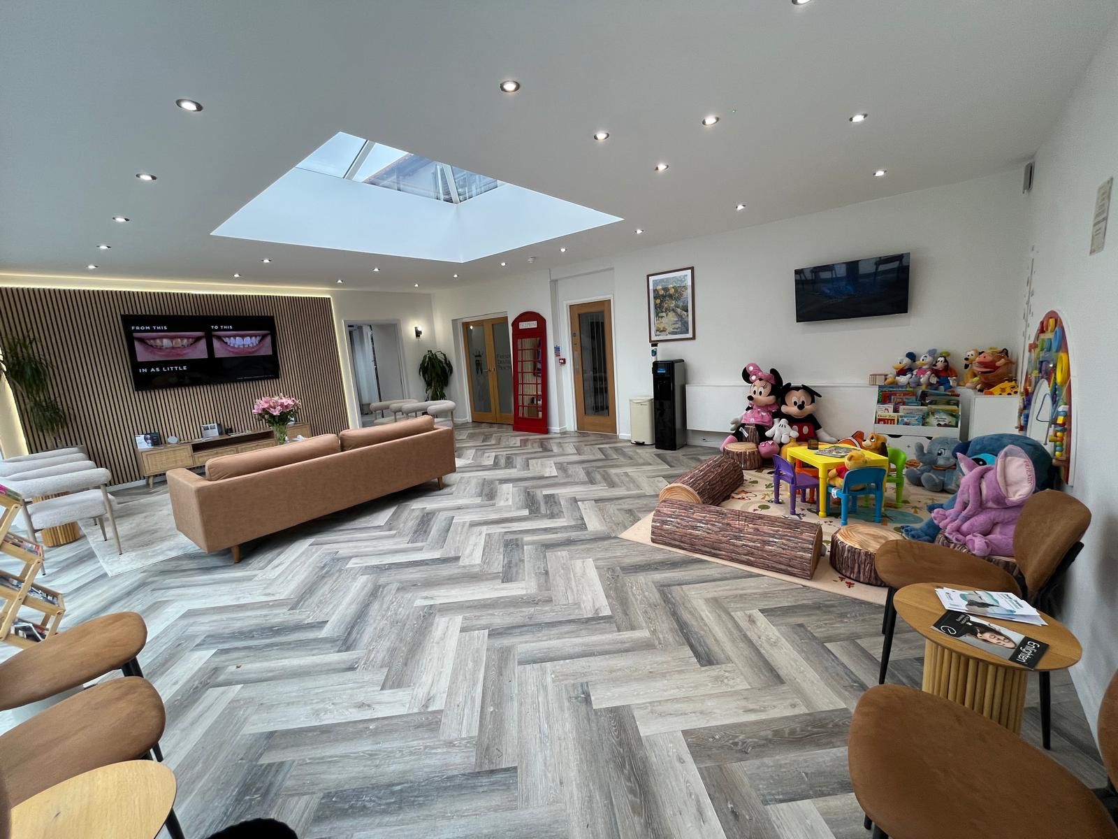 Modern spacious waiting room with grey herringbone floor, brown sofa, children's play area with toys, skylight, and wall-mounted TV screens.