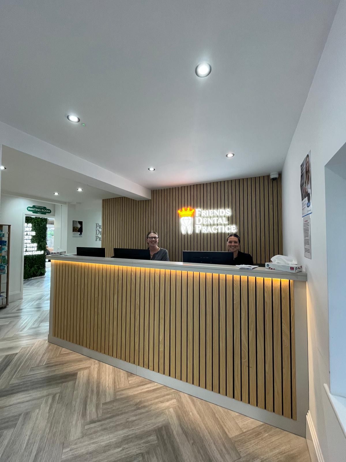 Reception desk with two smiling staff at Friends Dental Practice with wood paneling and illuminated sign behind.
