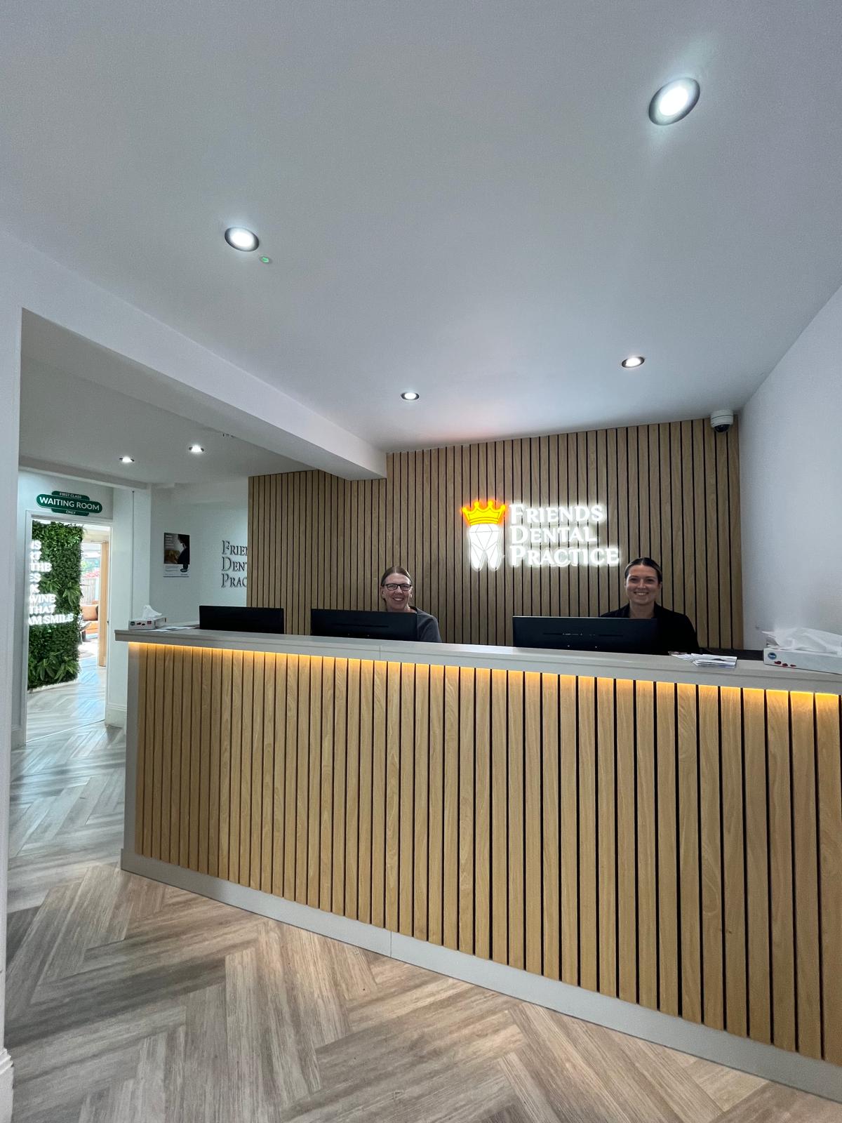 Reception desk with two smiling receptionists at Friends Dental Practice, featuring wood paneling and a lit logo on the wall.