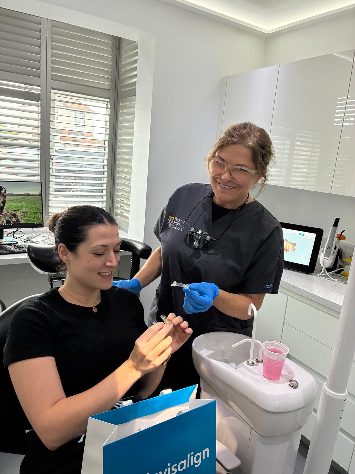 A dental professional in black scrubs shows Invisalign clear aligners to a seated female patient holding an aligner in a dental office.