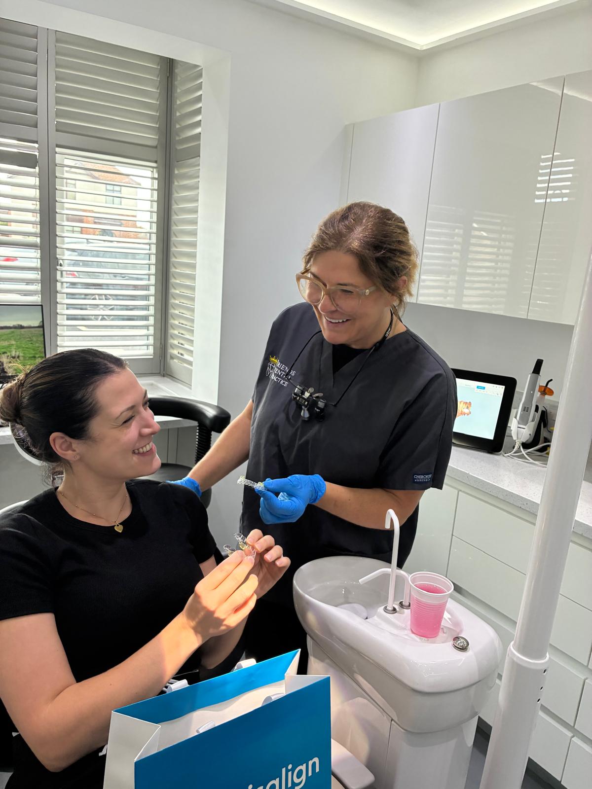 Dentist in black scrubs smiling and holding clear dental aligners while talking to a seated female patient who is also holding an aligner in a dental office.