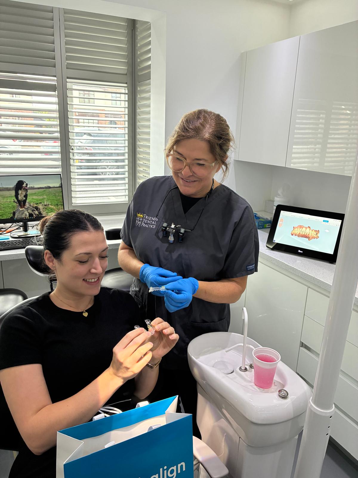 Dentist in gray scrubs with blue gloves showing a clear dental aligner to a smiling seated patient holding a similar aligner in a dental office.