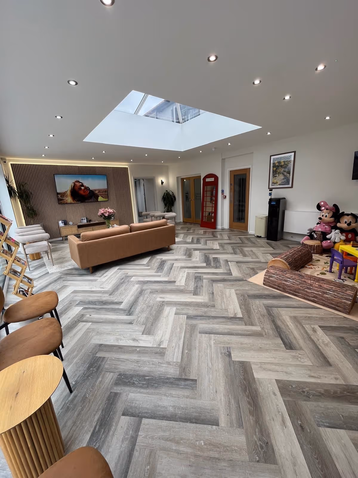 Modern waiting room with herringbone patterned floor, brown sofa, skylight, TV, red telephone booth, and children's play area with Mickey and Minnie Mouse plush toys.