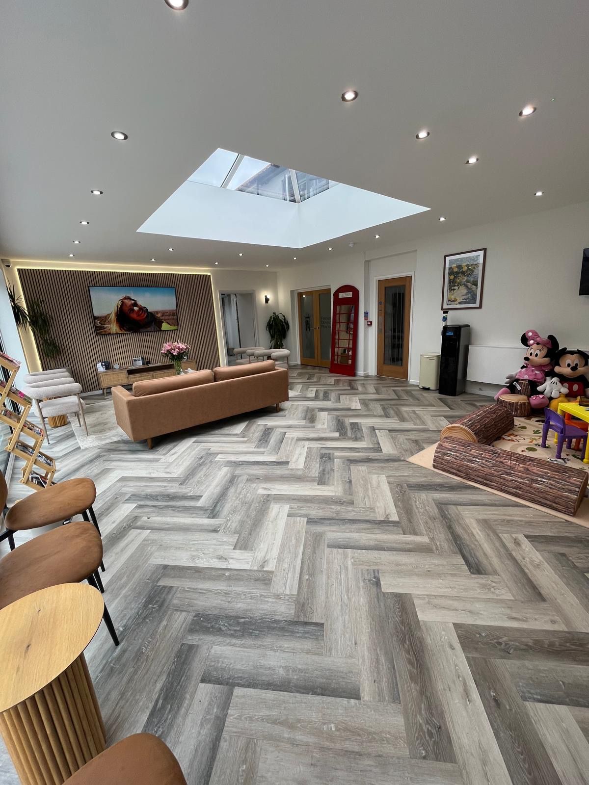 Modern waiting room with herringbone patterned floor, brown sofa, skylight, TV, red telephone booth, and children's play area with Mickey and Minnie Mouse plush toys.
