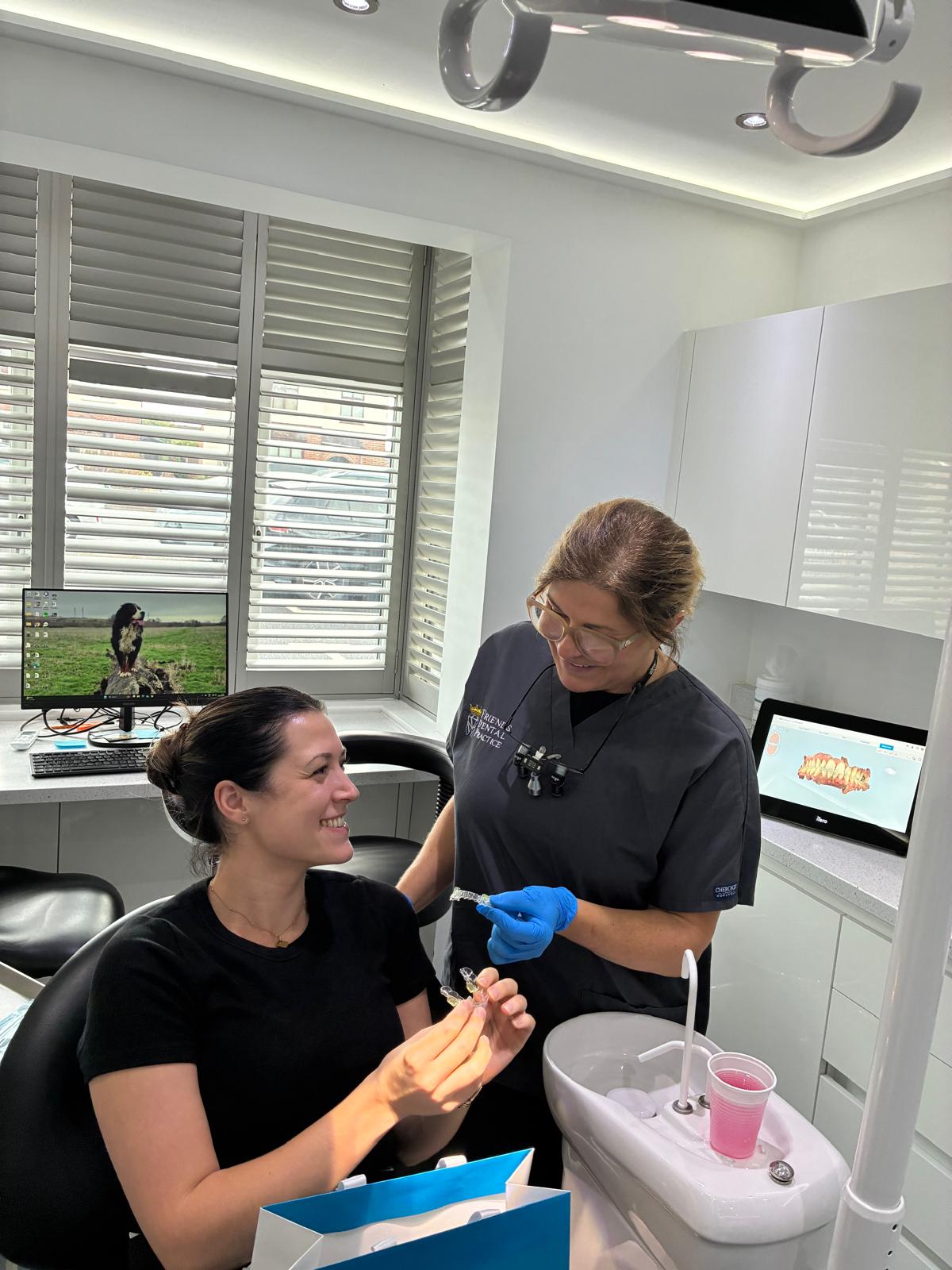 Dentist wearing scrubs and gloves showing clear dental aligners to a smiling patient in a bright dental clinic room.