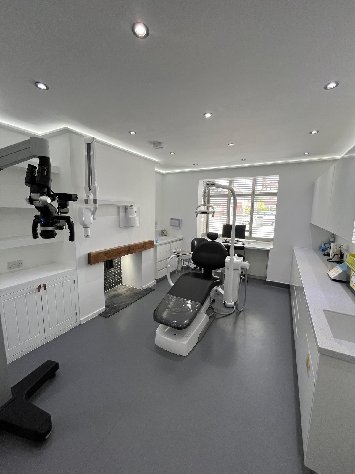 Modern dental clinic room with dental chair, overhead light, microscope, white cabinetry, and window with blinds.
