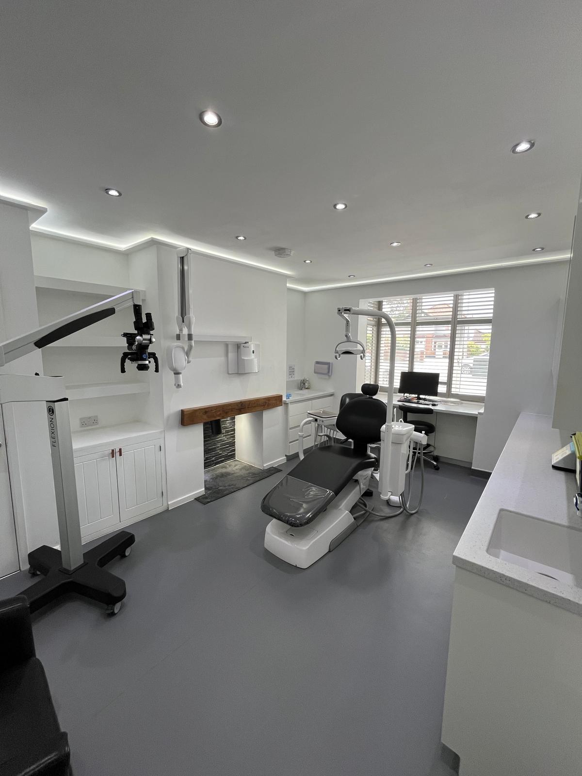 Modern dental office with black dental chair, overhead light, microscope on stand, computer on desk, and white cabinetry.