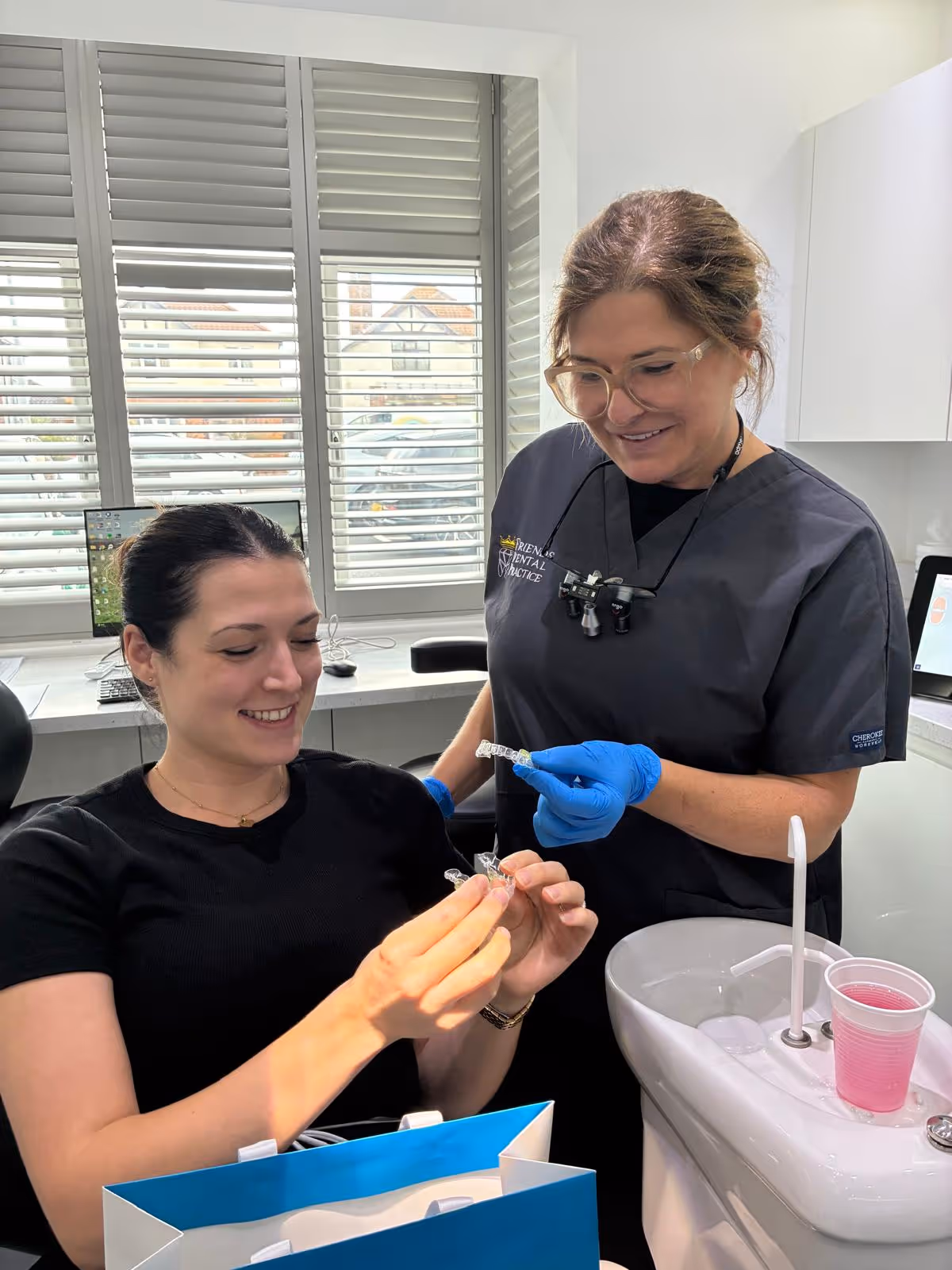 Dentist in black scrubs with magnifying glasses showing a clear dental aligner to a smiling patient sitting in a dental chair.