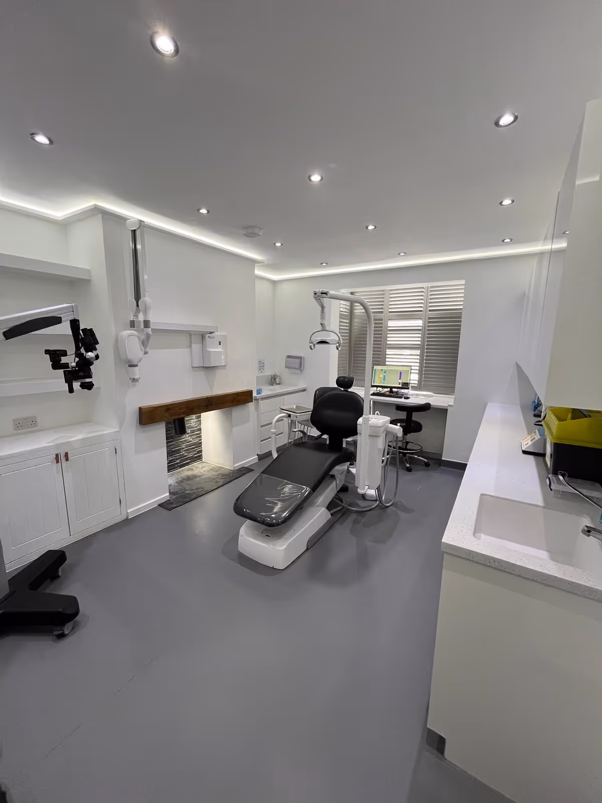 Modern dental clinic room with a black dental chair, overhead light, computer workstation, and white cabinets.
