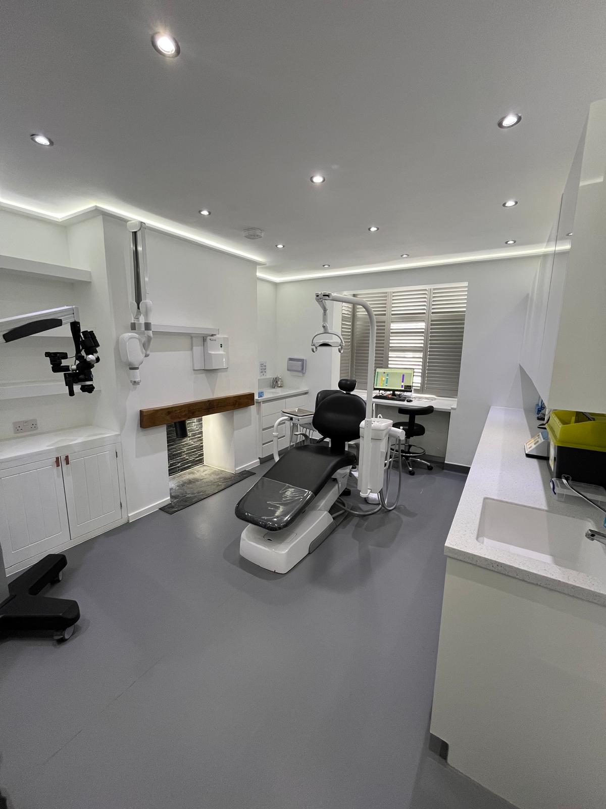 Modern dental clinic room with a black dental chair, overhead light, computer workstation, and white cabinets.