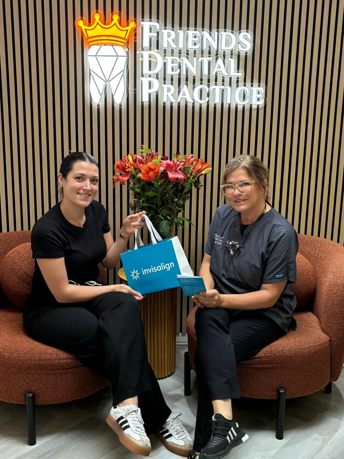 Two women sitting on brown chairs in a dental office waiting area with a 'Friends Dental Practice' sign and a flower arrangement behind them; one woman holds an Invisalign bag.
