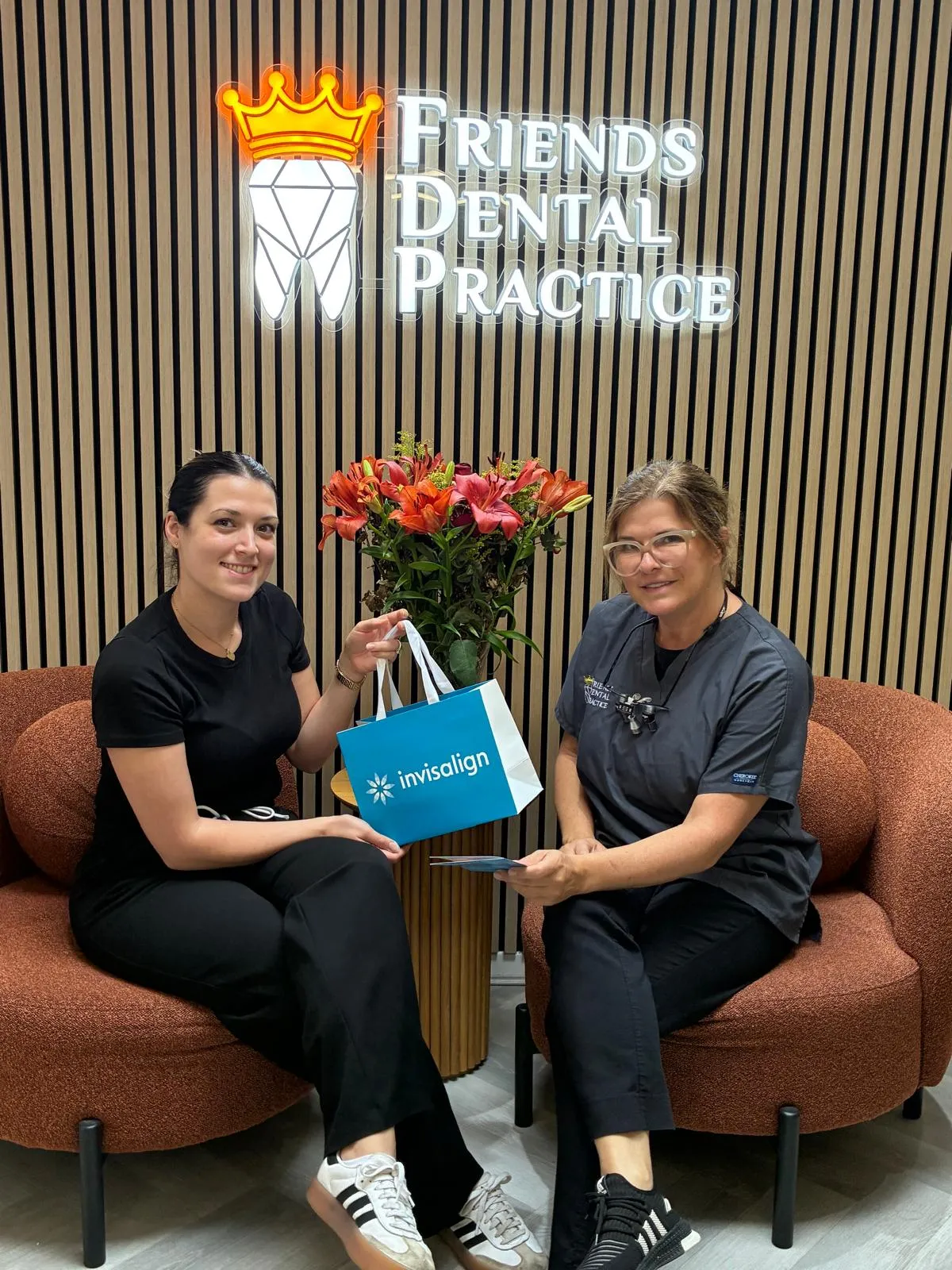 Two women sitting in a dental office waiting area with a 'Friends Dental Practice' sign behind them; one holds an Invisalign branded bag.