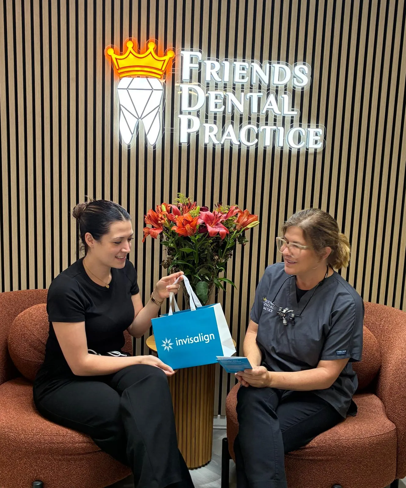 Two women sitting on chairs in a dental office under the 'Friends Dental Practice' sign, one holding an Invisalign bag.