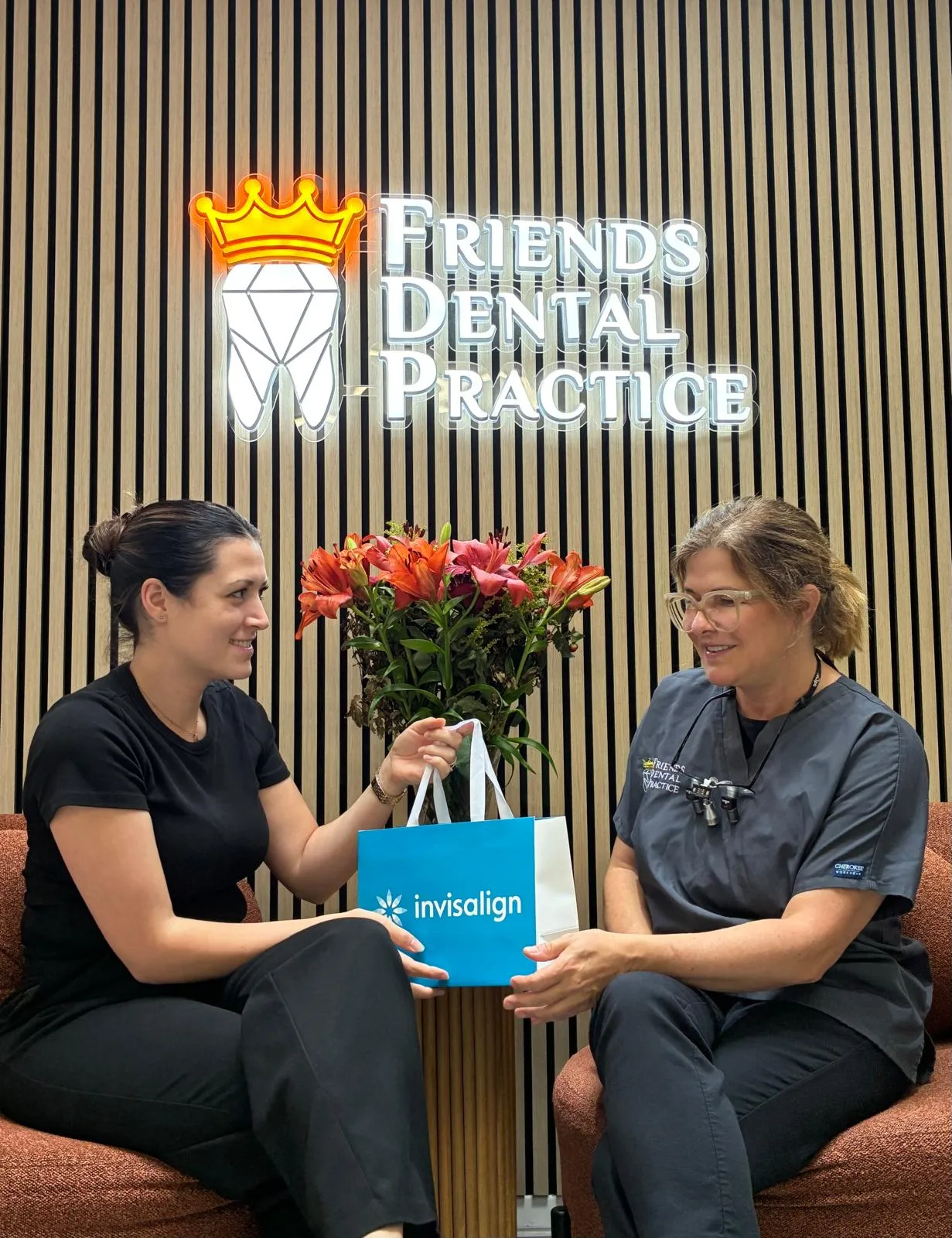 Two women sitting on chairs and exchanging a blue Invisalign bag in front of a wooden wall with a neon Friends Dental Practice sign and a vase of red flowers.