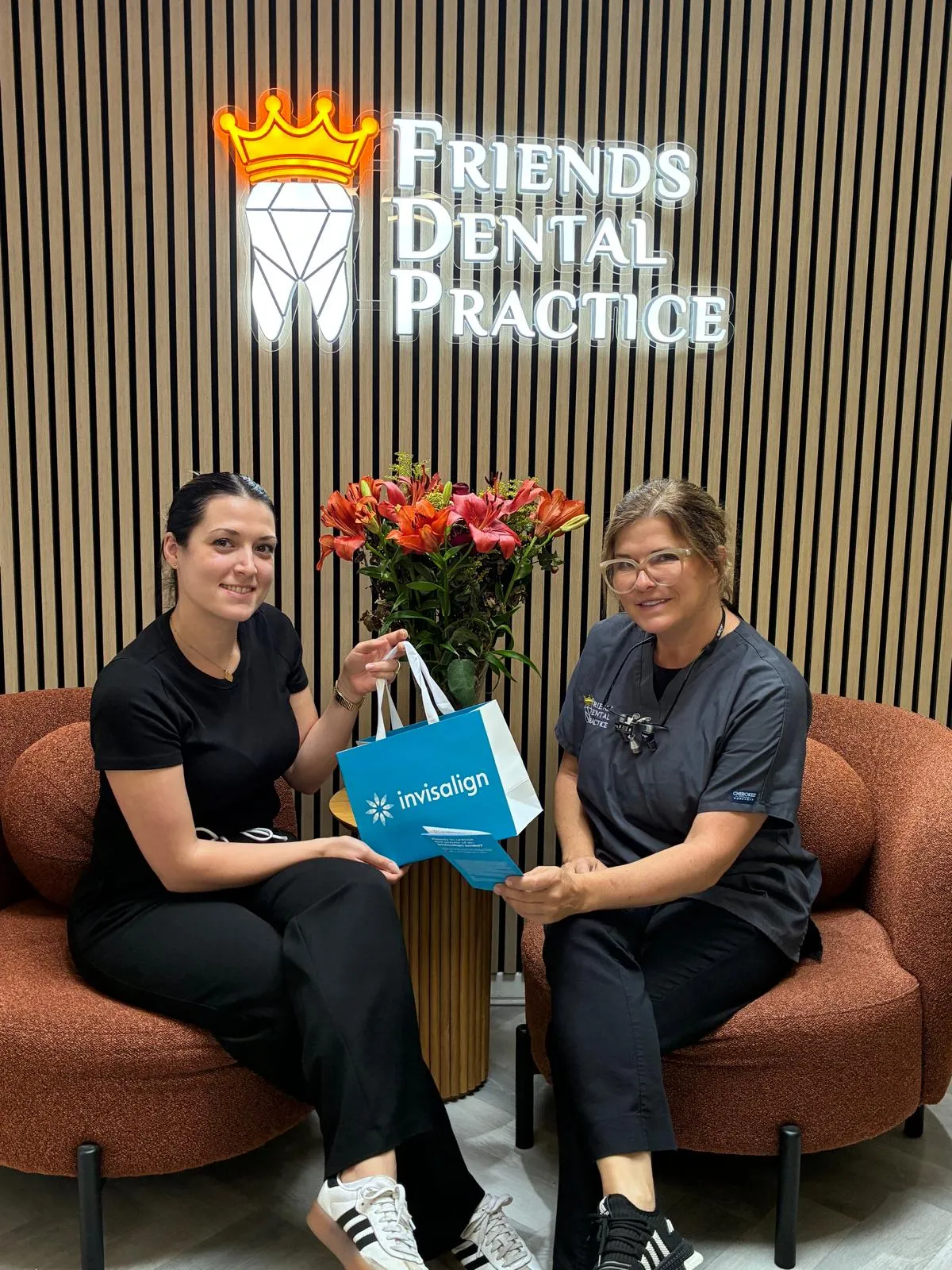 Two women sitting on brown chairs in front of a 'Friends Dental Practice' sign, holding an Invisalign bag and brochure with a vase of orange flowers between them.