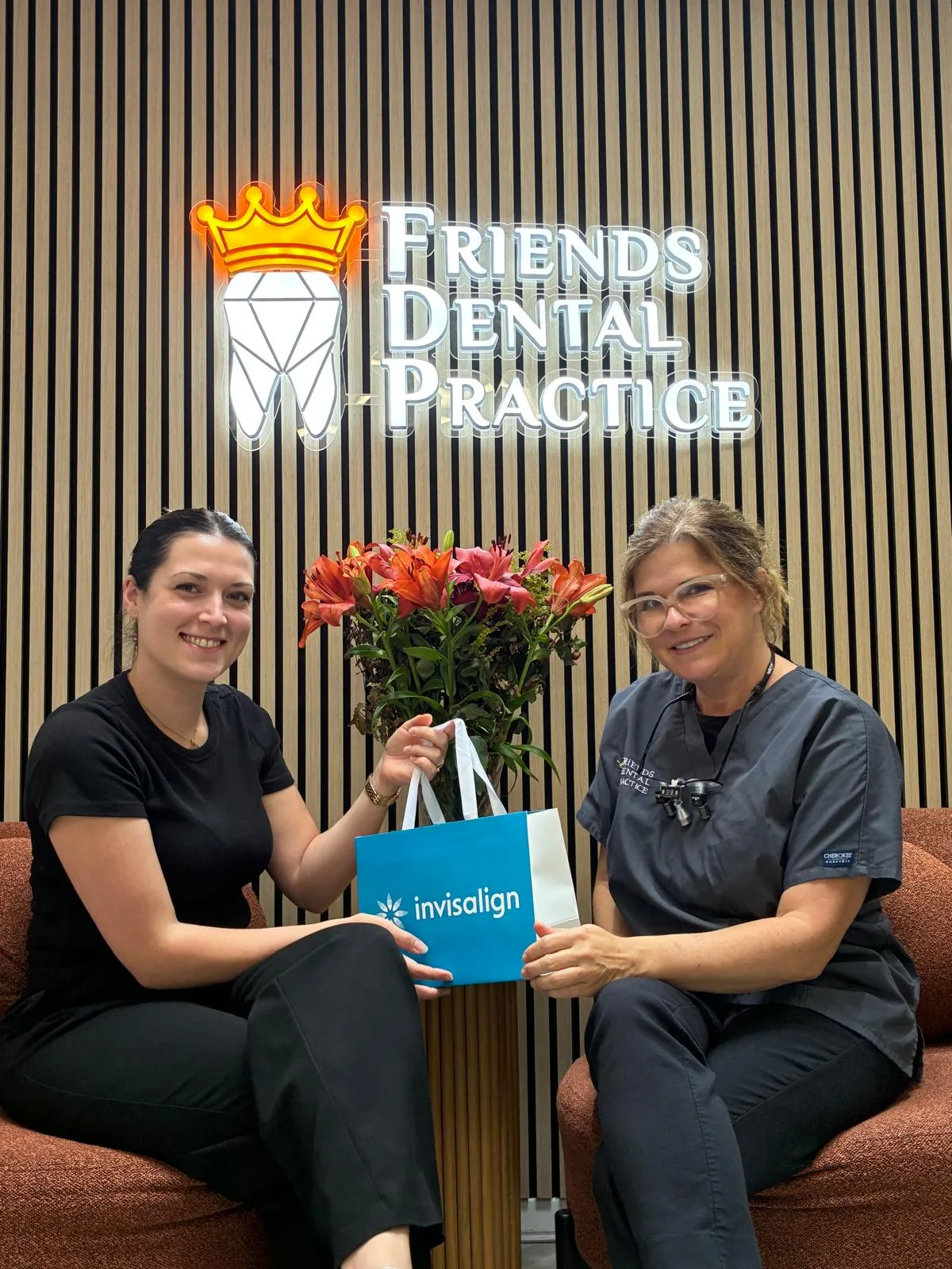 Two women sitting on chairs in a dental office holding a blue Invisalign bag, with a sign reading Friends Dental Practice behind them.