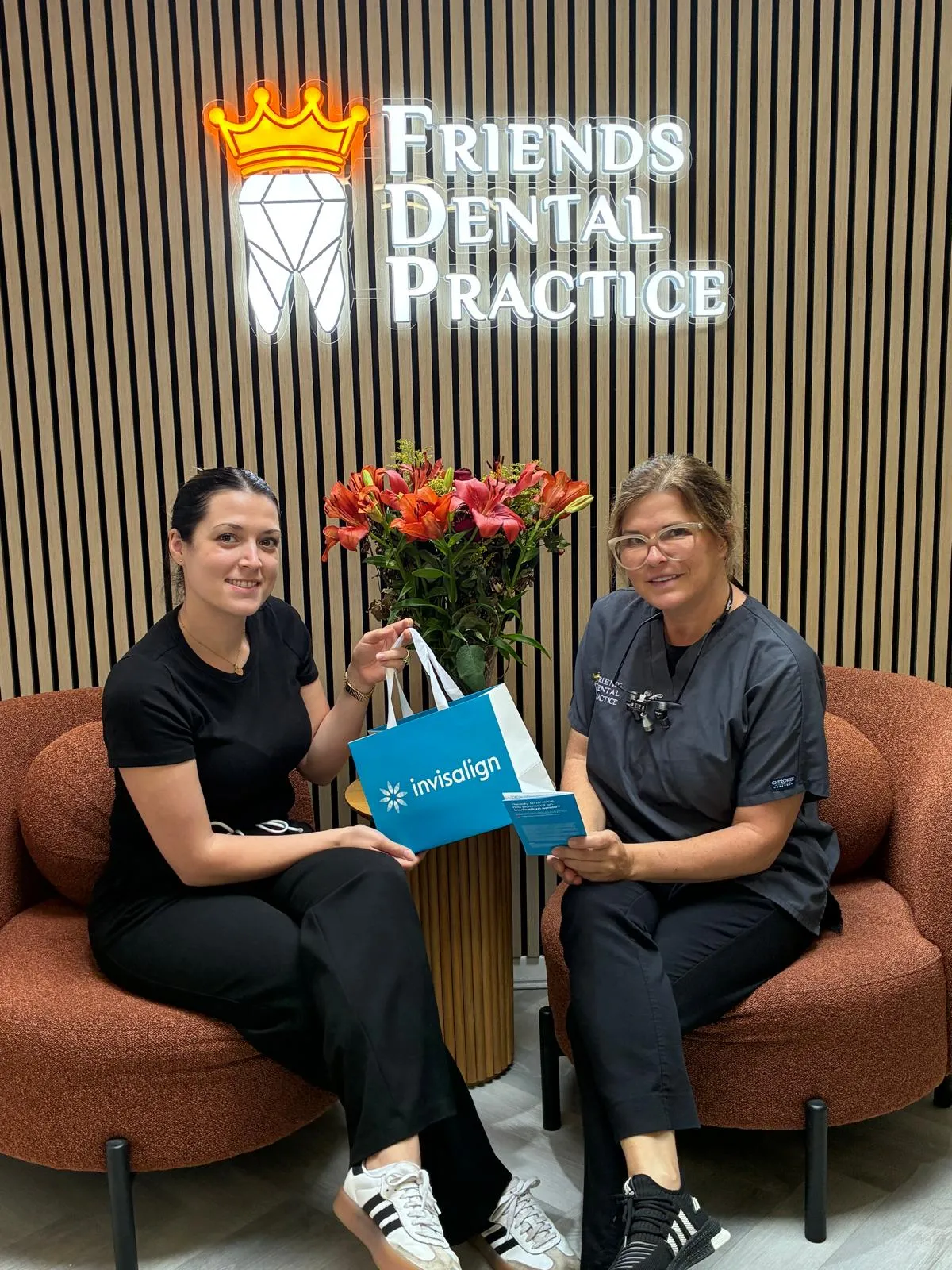 Two women seated on orange chairs at Friends Dental Practice, one holding an Invisalign bag and the other holding a pamphlet, with a flower arrangement between them.