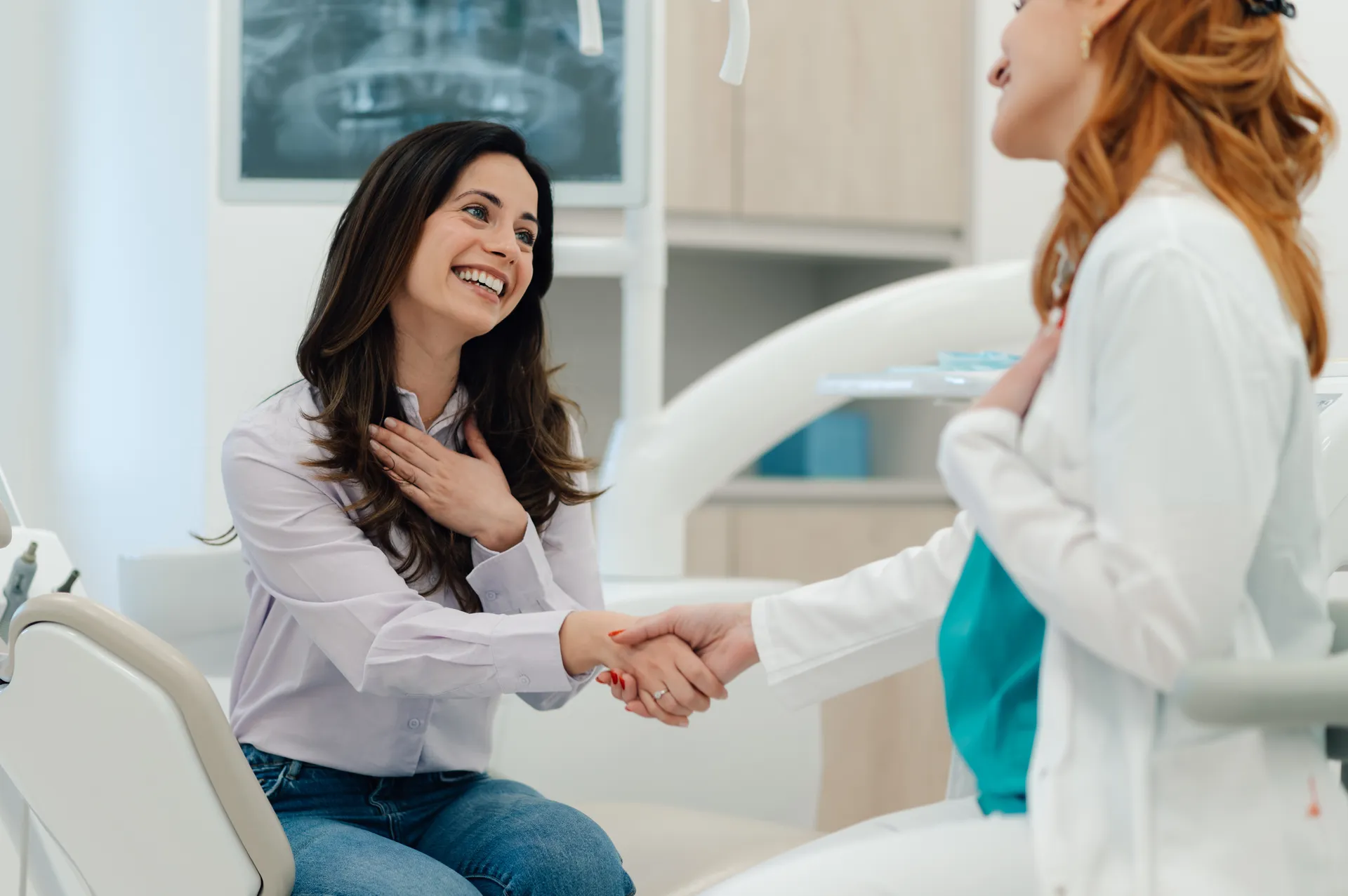 Smiling woman sitting in a dental chair shakes hands with a female dentist in a white coat in a dental clinic.