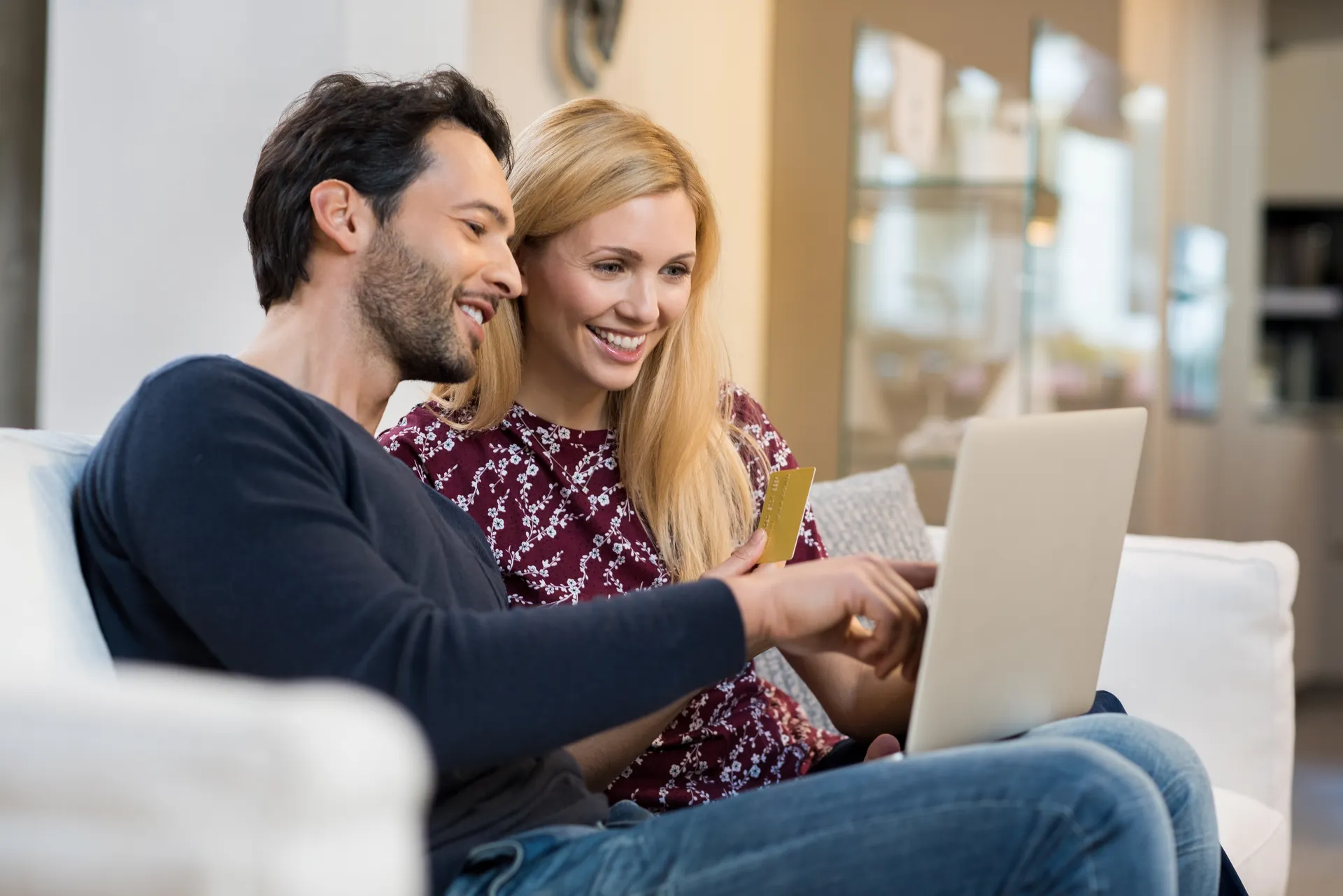 Smiling couple sitting on a couch using a laptop and holding a credit card, shopping online.