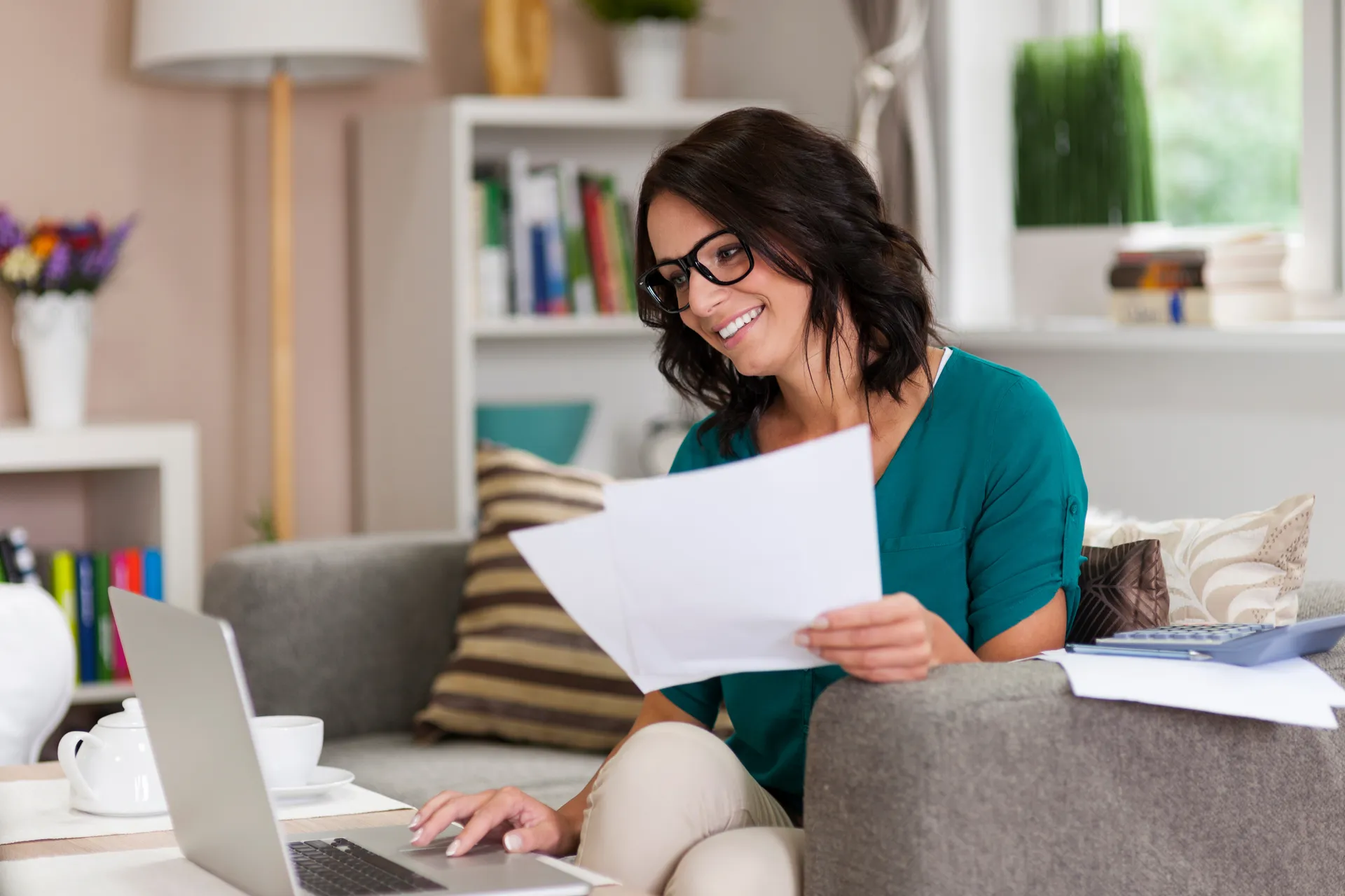 Smiling woman wearing glasses holding papers and using a laptop while sitting on a sofa in a cozy living room.