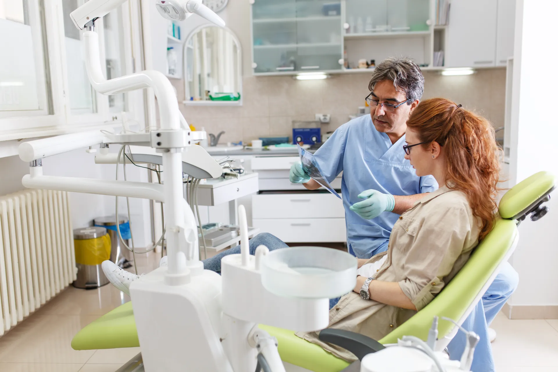 Dentist in scrubs showing a dental X-ray to a woman seated in a green dental chair inside a clinic.